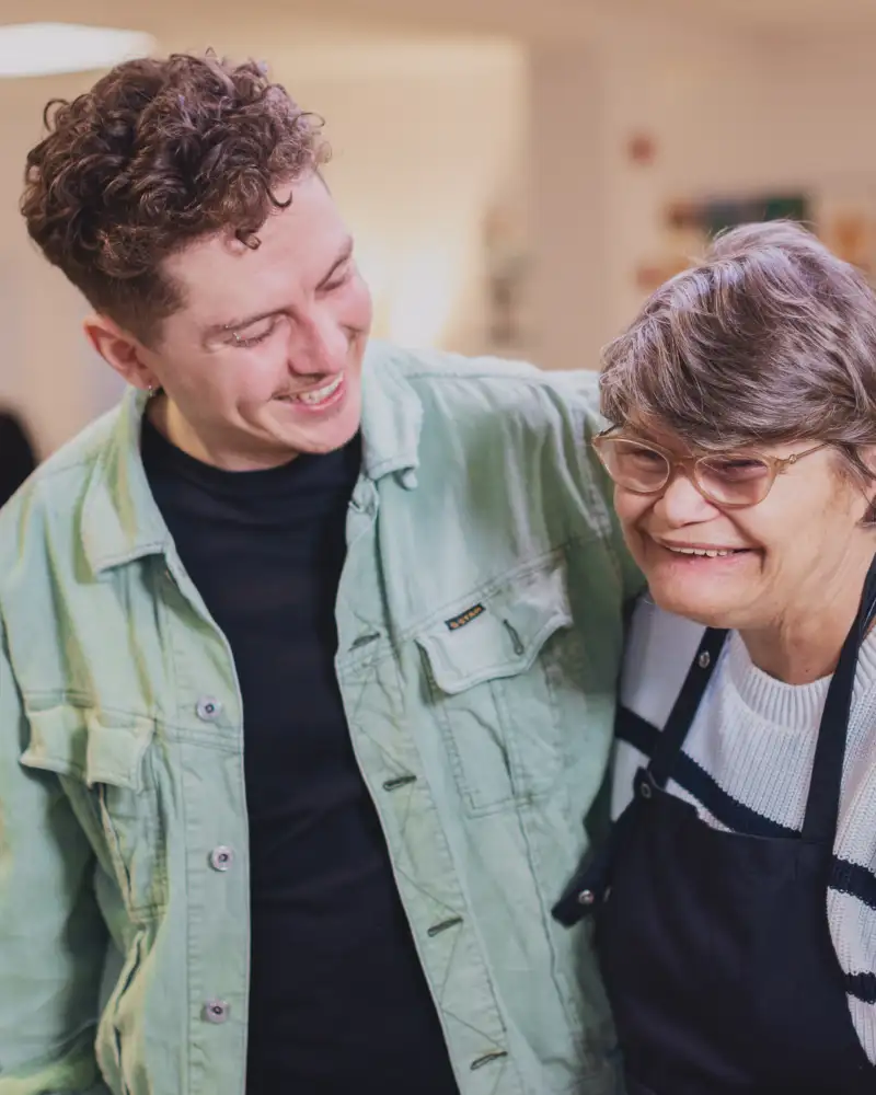 In einem Raum einer Begegnungsstätte: Zwei Menschen legen sich freundschaftlich die Arme um die Schultern und lachen. Der Linke ist ein junger Mann mit Jeansjacke und gewellten kurzen braunen Haaren. Die Rechte eine etwas kleinere, ältere Frau mit einer  Behinderung. Sie trägt eine Schürze über ihrem Pulli und hat eine Brille auf.
