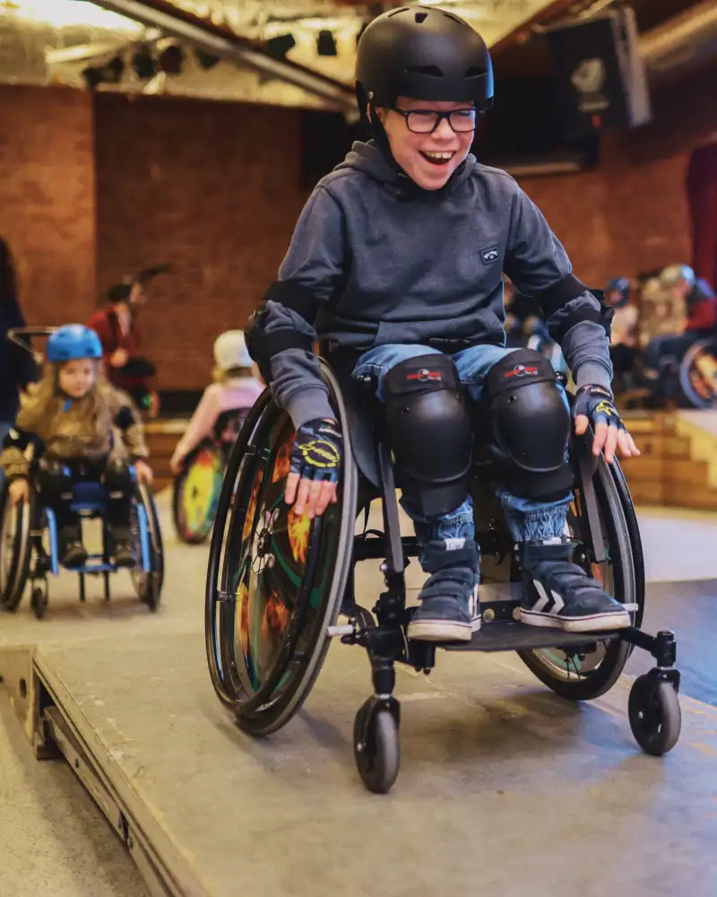 In einer Hall sind mehrere Rampen aufgebaut. Ein Junge fährt mit einem Sportrollstuhl über eine Rampe. Hinter ihm folgt ein kleines Mädchen. Beide tragen Helme. Dahinter steht einer Frau.