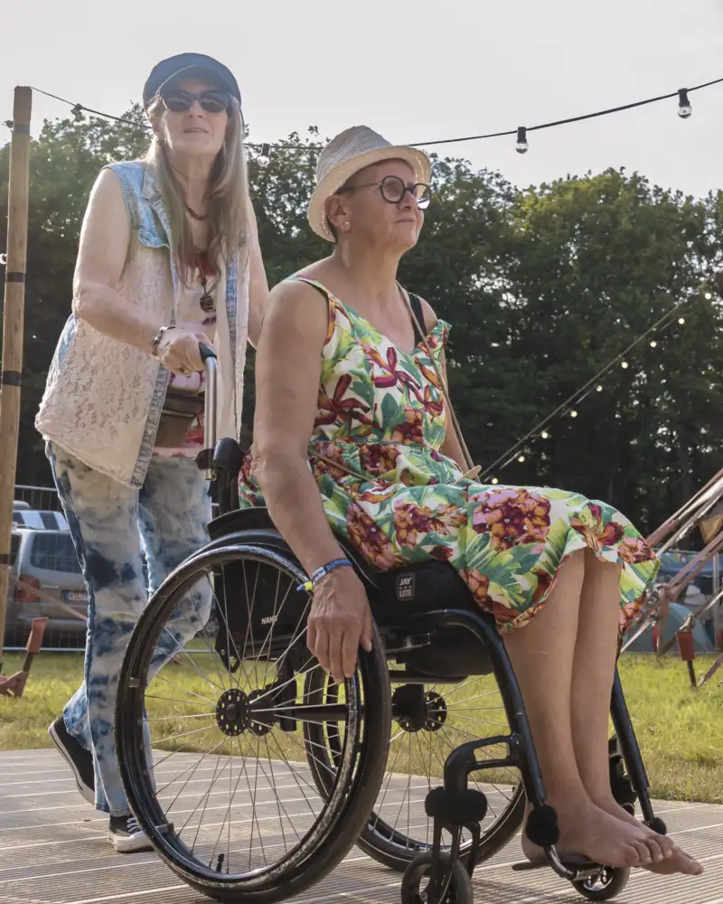 Besucher*innen auf einem Festival. Eine Frau im Sommerkleid und Sonnenhut wird von einer anderen Frau im Rollstuhl geschoben.