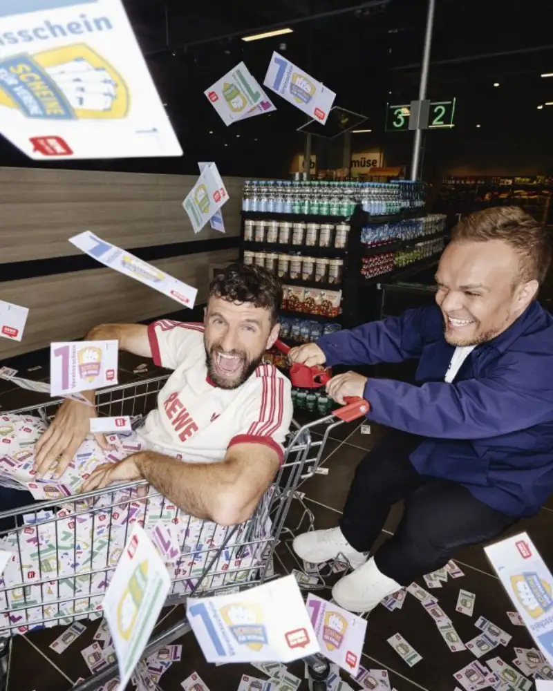 Fußballer Thomas Müller sitzt in einem Einkaufswagen, der vom Para-Sportler Niko Kappel geschoben wird. Beide lachen und um sie herum fliegen Vereinsscheine von REWE.
