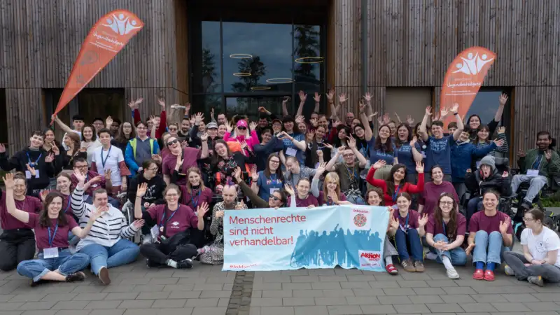 Eine Gruppe von etwa 80 Jugendlichen und jungen Erwachsenen mit und ohne Behinderung posiert vor dem Eingang eines holzgetäfelten Tagungshauses für ein Gruppenbild. Die in der ersten Reihe sitzenden Teilnehmer*innen halten ein 2-Meter-langes Banner mit der Aufschrift "Menschenrechte sind nicht verhandelbar". Hinter der Gruppe stehen Beachflags mit dem Logo der Servicestelle Jugendbeteiligung.