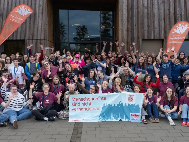 Eine Gruppe von etwa 80 Jugendlichen und jungen Erwachsenen mit und ohne Behinderung posiert vor dem Eingang eines holzgetäfelten Tagungshauses für ein Gruppenbild. Die in der ersten Reihe sitzenden Teilnehmer*innen halten ein 2-Meter-langes Banner mit der Aufschrift "Menschenrechte sind nicht verhandelbar". Hinter der Gruppe stehen Beachflags mit dem Logo der Servicestelle Jugendbeteiligung.