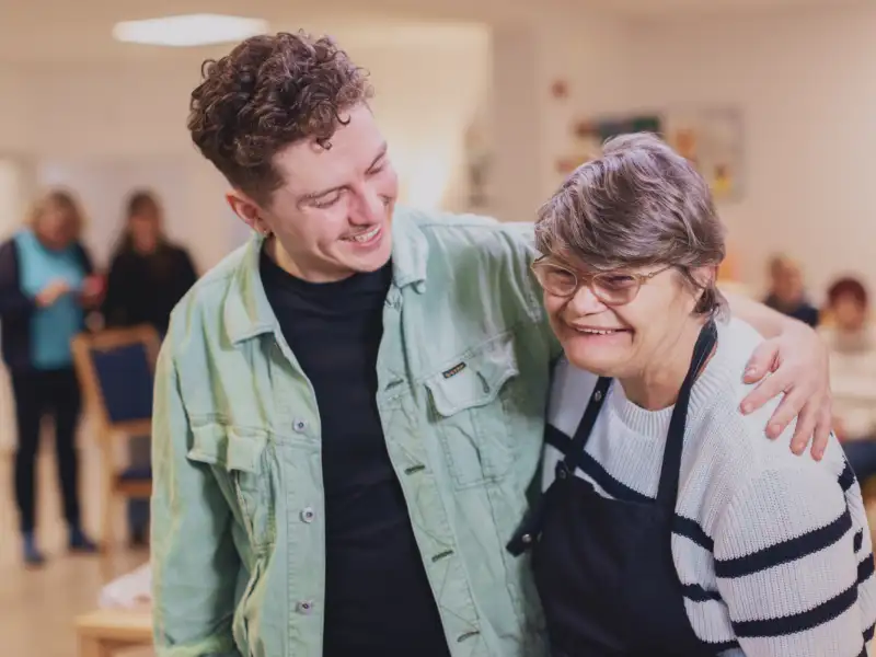 In einem Raum einer Begegnungsstätte: Zwei Menschen legen sich freundschaftlich die Arme um die Schultern und lachen. Der Linke ist ein junger Mann mit Jeansjacke und gewellten kurzen braunen Haaren. Die Rechte eine etwas kleinere, ältere Frau mit einer  Behinderung. Sie trägt eine Schürze über ihrem Pulli und hat eine Brille auf.