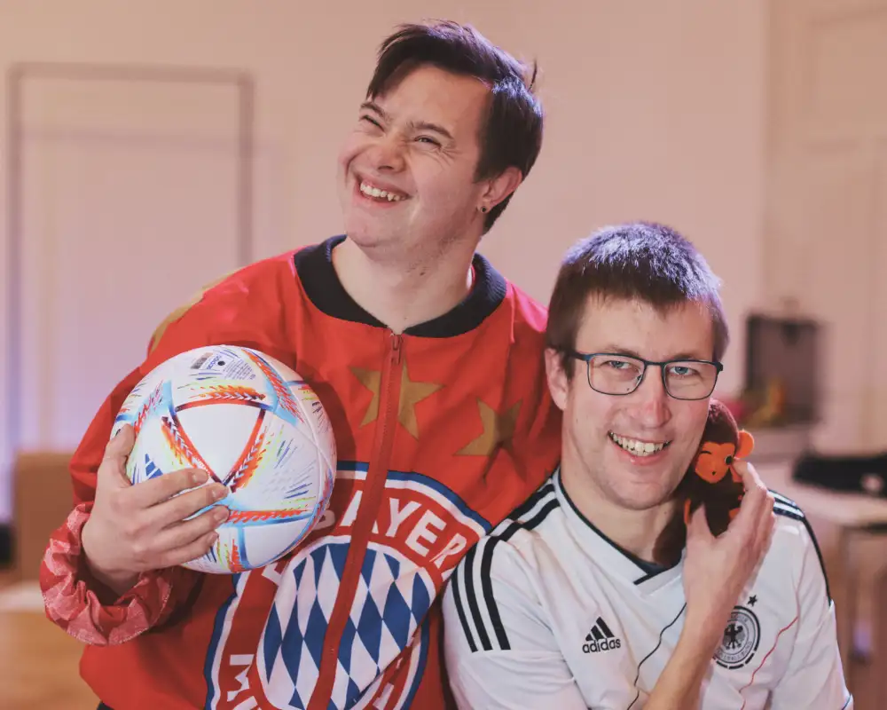 Zwei junge Männer in Sporttrikots stehen lachend nebeneinander. Der eine hält eine Fußball unter dem Arm.