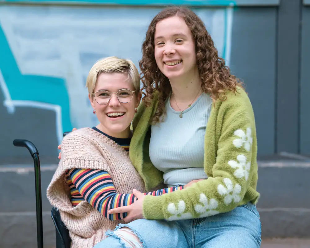 Zwei junge Frauen umarmen sich freundschaftlich. Eine von ihnen sitzt im Rollstuhl.