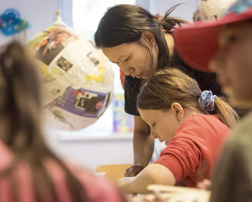 Ein Mädchen und eine junge Frau beugen sich gemeinsam über einen Tisch. Im Vordergrund sieht man unscharf zwei weitere Kinder. Im Hintergrund erkennt man ein Kunstobjekt.