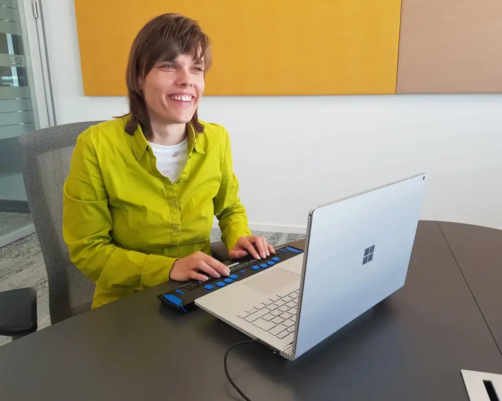 Eine blinde junge Frau sitzt lächelnd vor einem Laptop mit Braillezeile.