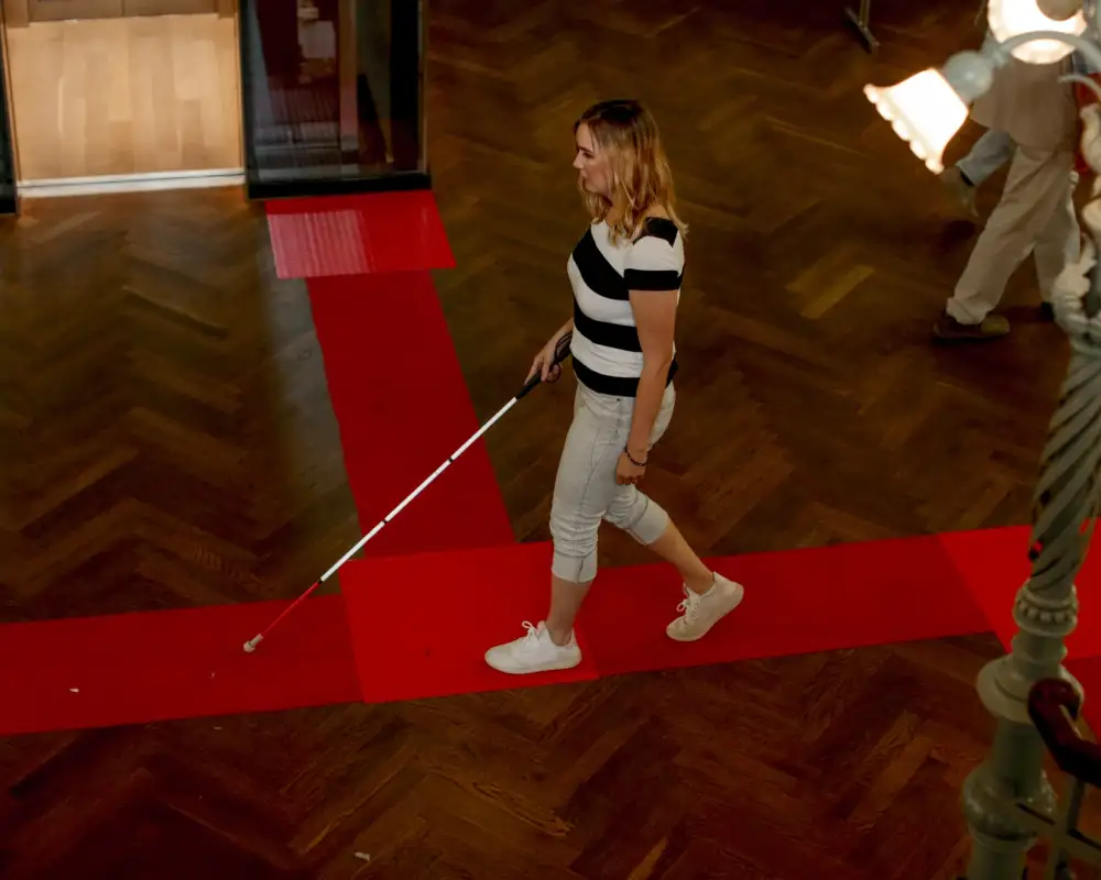 In einem großen Raum geht eine Frau mit einem Blindenlangstock an einer Markierung entlang.