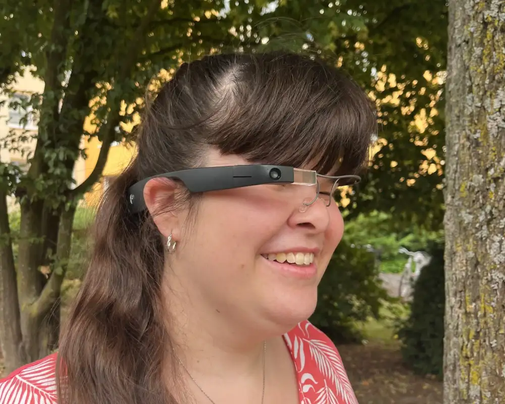 Junge Frau mit langen dunklen Haaren und sommerlicher roter Bluse trägt ein Brillengestell, in dessen rechten Bügel eine Kamera, ein kleines Display und weitere Technik integriert sind.