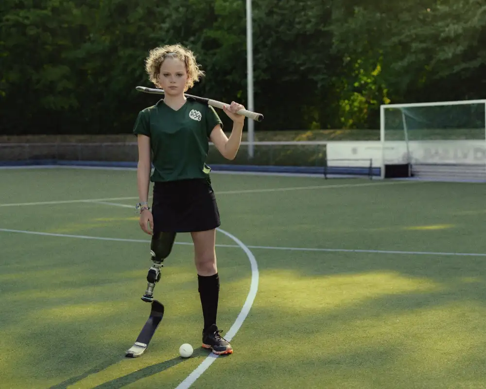Annika, ein Mädchen mit lockigem Haar und Beinprothese steht auf einem Hockeyplatz. Zwischen ihren Beinen liegt der Ball, ihren Schläger hat sie lässig über die Schultern gelegt.