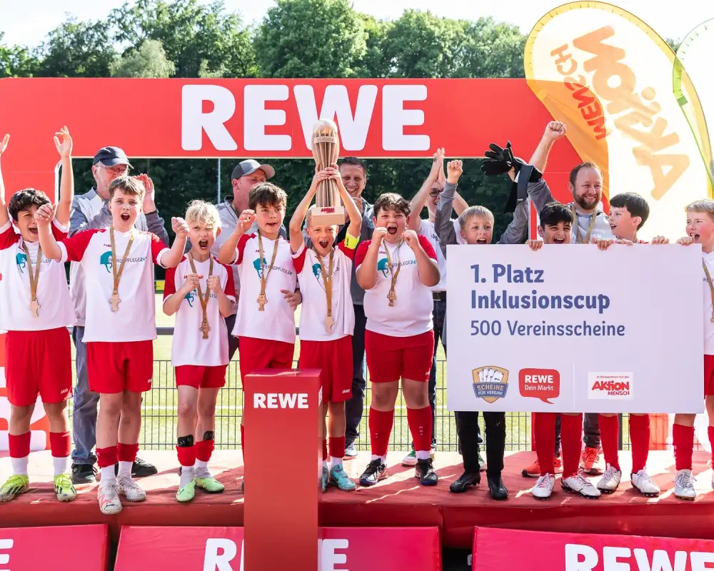Eine Jungen-Fußballmannschaft feiert auf einem Podest bei der Siegerehrung ihren 1. Platz. Der Kapitän in der Mitte hält den Pokal in die Höhe, drei andere Jungen (etwa 12 Jahre) halten ein Schild in den Händen auf dem steht: "1. Platz Inklusionscup - 500 Vereinsscheine".