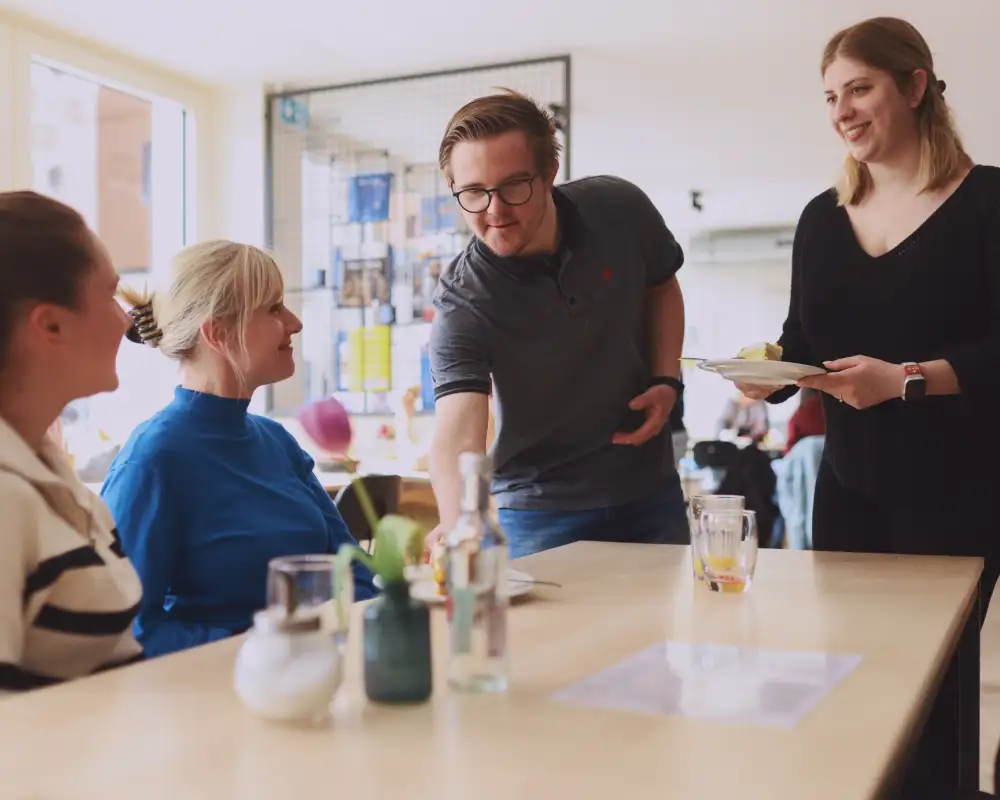 Zwei Frauen sitzen an einem Holztisch. Ein Mann mit Down-Syndrom und eine junge Frau servieren jeder einen Teller mit Kuchen.