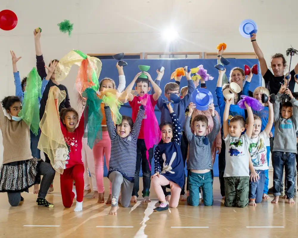 Eine Gruppe von Kindern in einer Turnhalle. Alle jubeln und halten bunte Tücher, Diabolos und Bälle in die Luft.