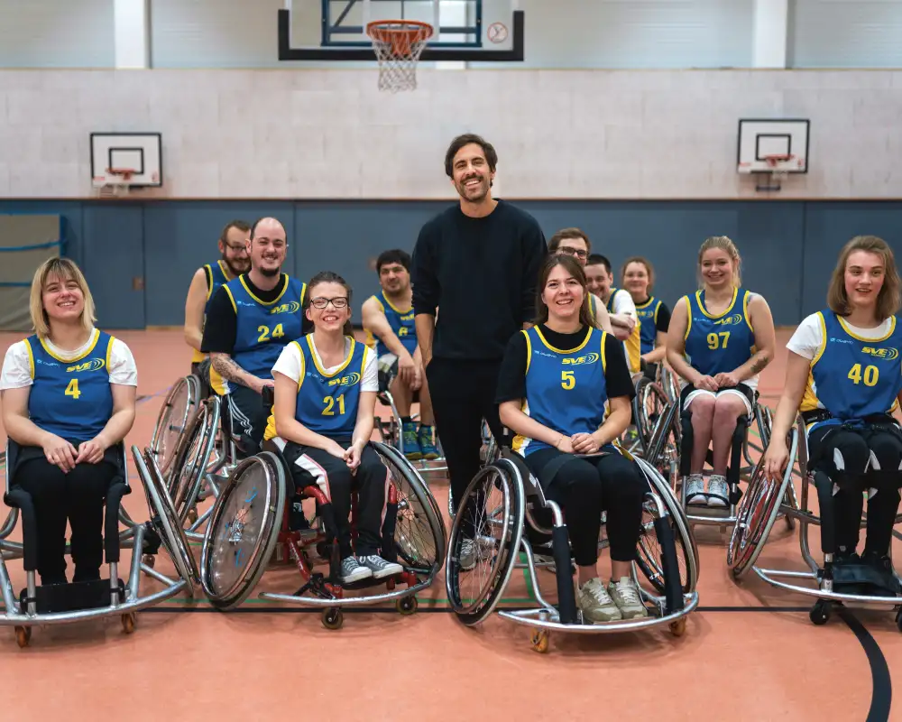 Max Giesinger steht in einer Turnhalle in mitten einer Gruppe Rollstuhl-Basketballer*innen