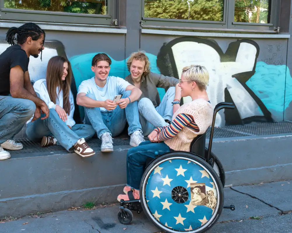 5 gut gelaunte Jugendliche. Sie haben unterschiedliche Hautfarbe. Eine von ihnen sitzt im Rollstuhl.