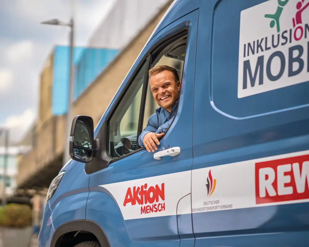 In einem blauen Kleintransporter sitzt ein kleinwüchsiger, sportlicher Mann mit kurzen blonden Haaren. Er sieht aus dem Fenster der Fahrertür und lacht in die Kamera. Auf dem Bus sind Ausfkleber angebracht. Auf einem steht "Inklusionsmobil". Auf den anderen sind die Logos der kooperatiosnpartner zu sehen, die diesen Bus unterstützen: Aktion Mensch, Deutscher Behindertensportverband und REWE.