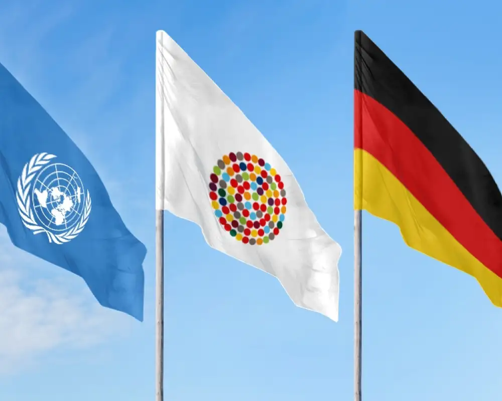 Drei Flaggen an Mast vor blauem Himmel: Eine mit dem Logo der UN, eine mit vielen bunten Punkten im Kreis, die Inklusion symbolisieren sollen und die deutsche Fahne