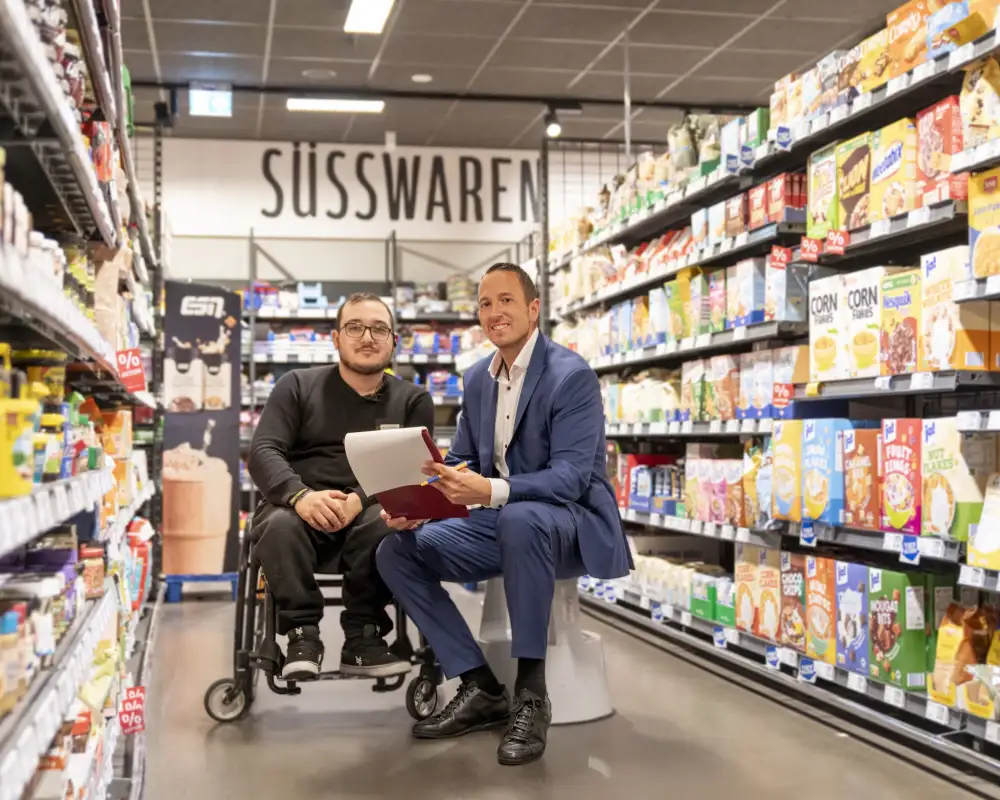 In einem Supermarkt-Gang zwischen zwei Regalreihen sitzen zwei Männer. Links ein Mann in schwarzer Hose und Pulli im Rollstuhl mit Brille und mit Bart, rechts neben ihm ein Mann mit kurzen dunklen Haaren in blauem Anzug und weißem Hemd auf einem Tritthocker. Beide lächeln in die Kamera.