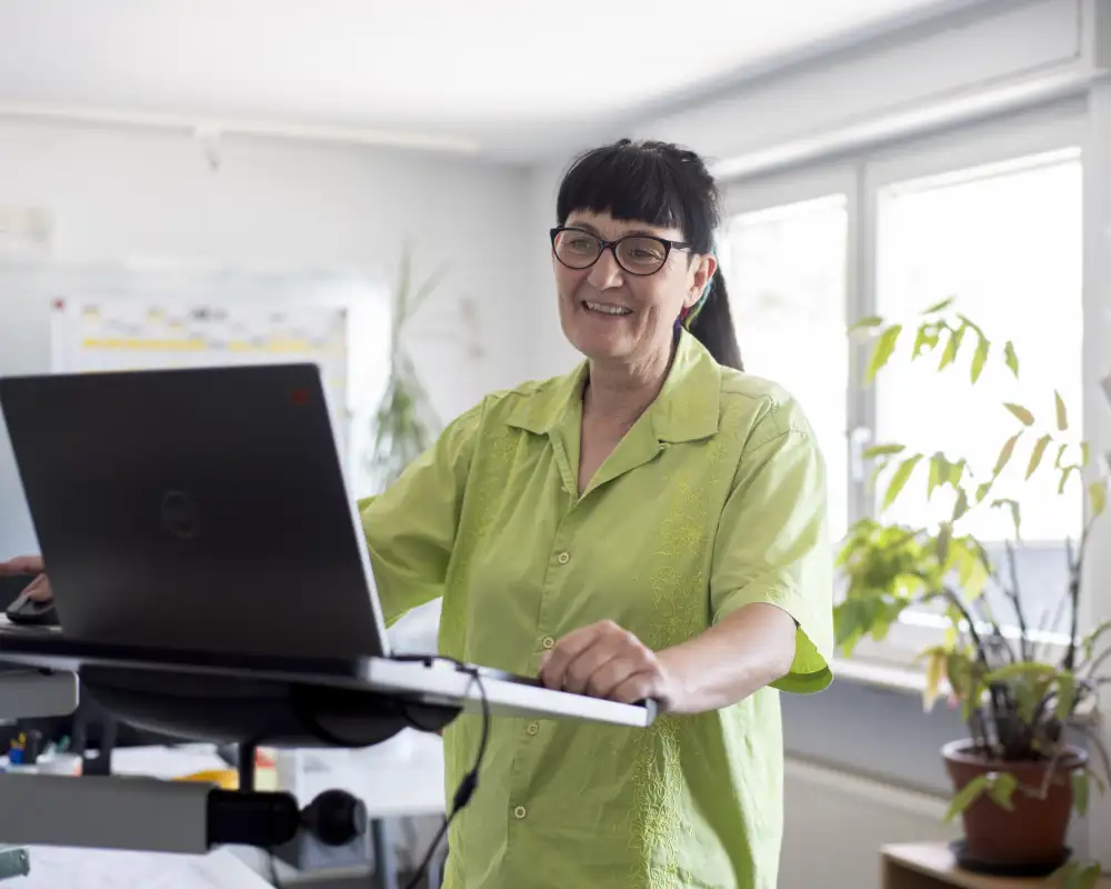 Eine Frau steht in einem Büroraum an einem Laptop. Sie trägt einen Brille, einen Pferdeschwanz und eine hellgrüne, kurzärmelige Bluse.