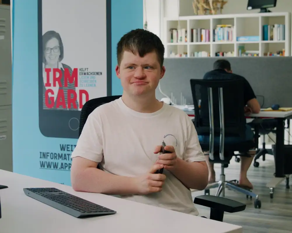 Ein junger Mann mit Behinderung sitzt in einem Büro am Schreibtisch. Er trägt ein weißes T-Shirt und schaut auf einen Computer-Monitor.