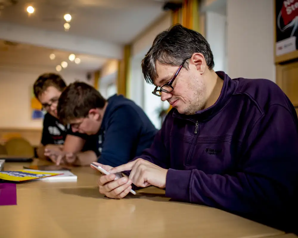 Ein Mann sitz an einem Tisch und schaut auf ein Smartphone. Neben ihm am Tisch sitzen zwei weitere Männer, die gemeinsam auf ein weiteres Smartphone gucken.