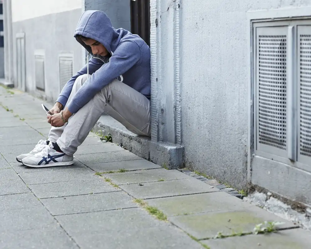 in Jugendlicher sitzt in einem Hauseingang. Er hat sich die Kapuze seines Pullis über den Kopf gezogen und schaut auf sein Handy.