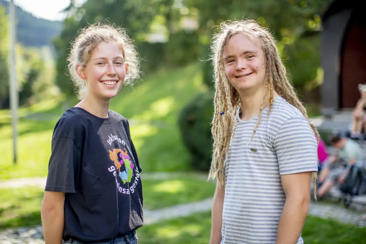 Zwei junge Erwachsene stehen sich in einem Park gegenüber. Ihre Köpfe sind Richtung Kamera gedreht. Beide lächeln in die Kamera.