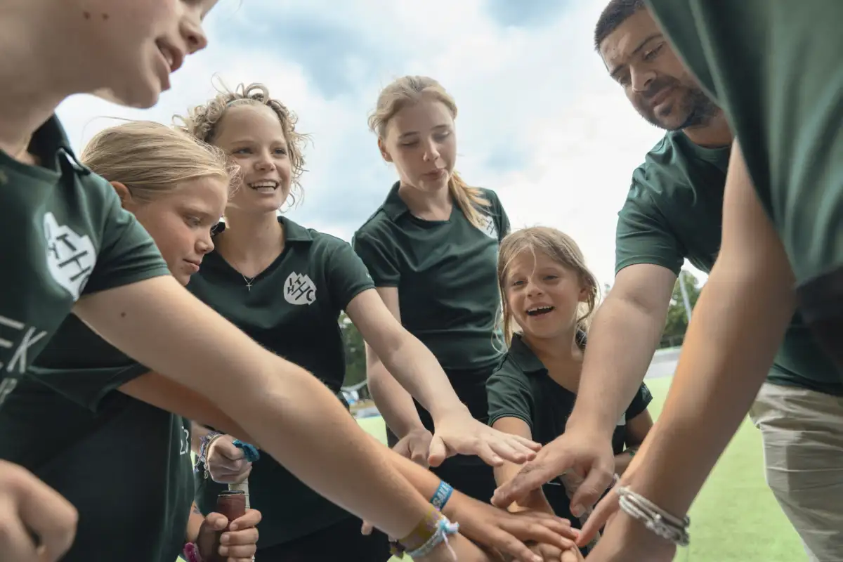 Eine Gruppe von Mädchen in grünen Sporttrikots mit ihrem Trainer. Sie schwören sich als Team ein, indem sie eine Runde bilden und alle ihre Hände aufeinandergelegt haben. Sie lächeln.