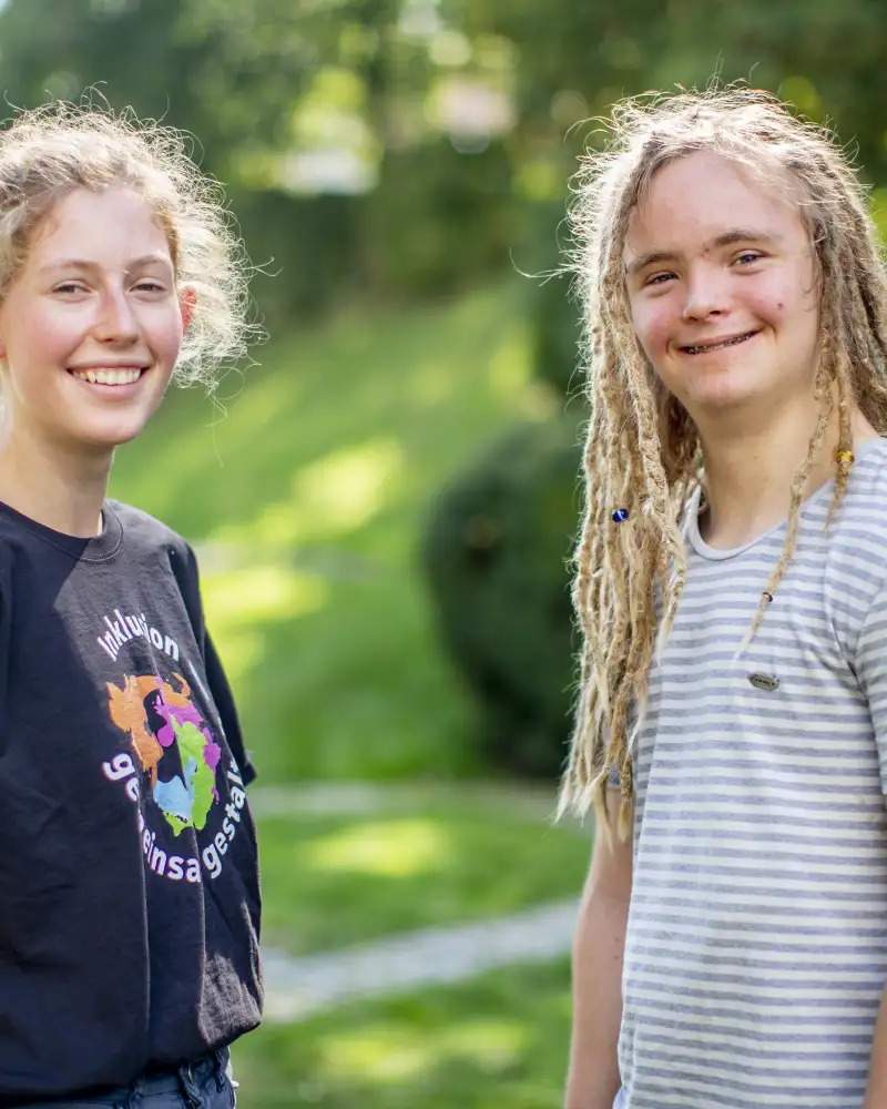 Zwei junge Erwachsene stehen sich in einem Park gegenüber. Ihre Köpfe sind Richtung Kamera gedreht. Beide lächeln in die Kamera.