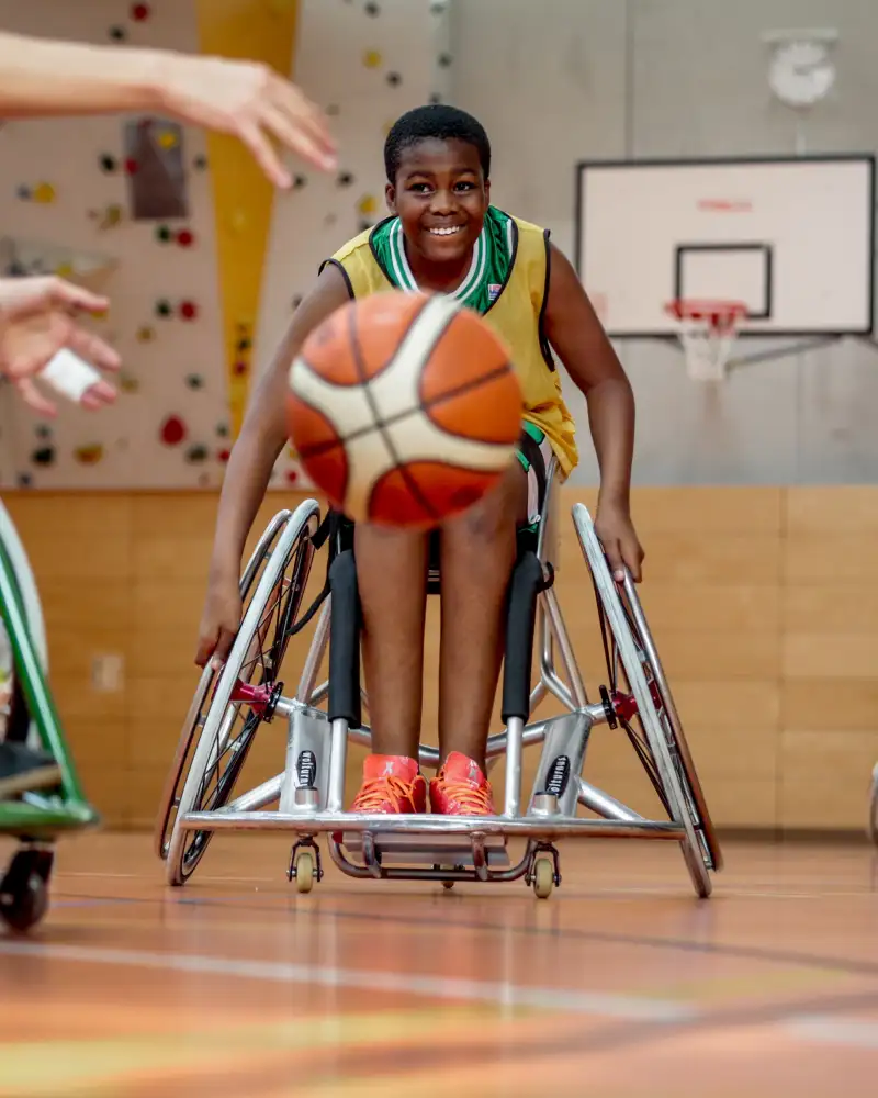 Ein Junge in einem Sportrollstuhl in einer Turnhalle. Er fährt auf einen Basketball zu. Von links sieht man die Beine und Arme einer weiteren Person im Rollstuhl, die Richtung Basketball greift.