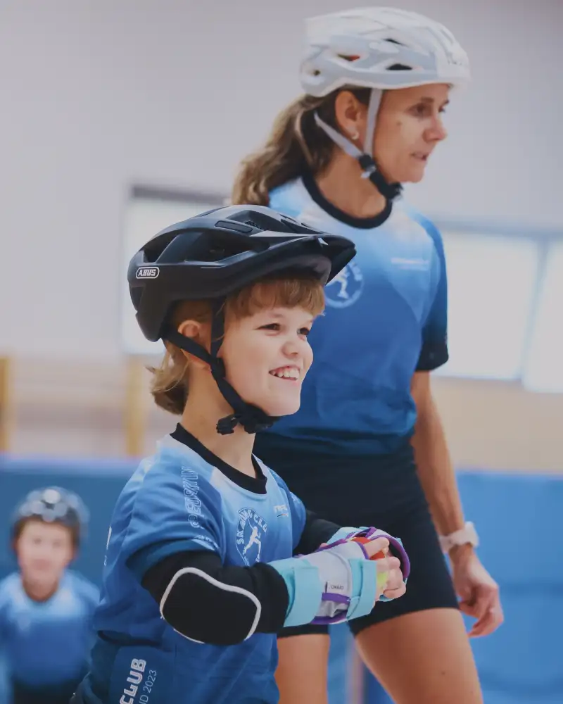 Zwei junge Frauen in einer Turnhalle. Sie tragen einen Fahrradhelm und ein Sportdress. Eine von beiden ist kleinwüchsig.