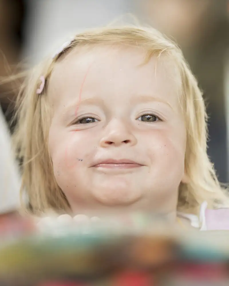 Ein kleines blondes Mädchen mit Spange im Haar schaut neugierig über einen Tisch hinweg direkt in die Kamera.