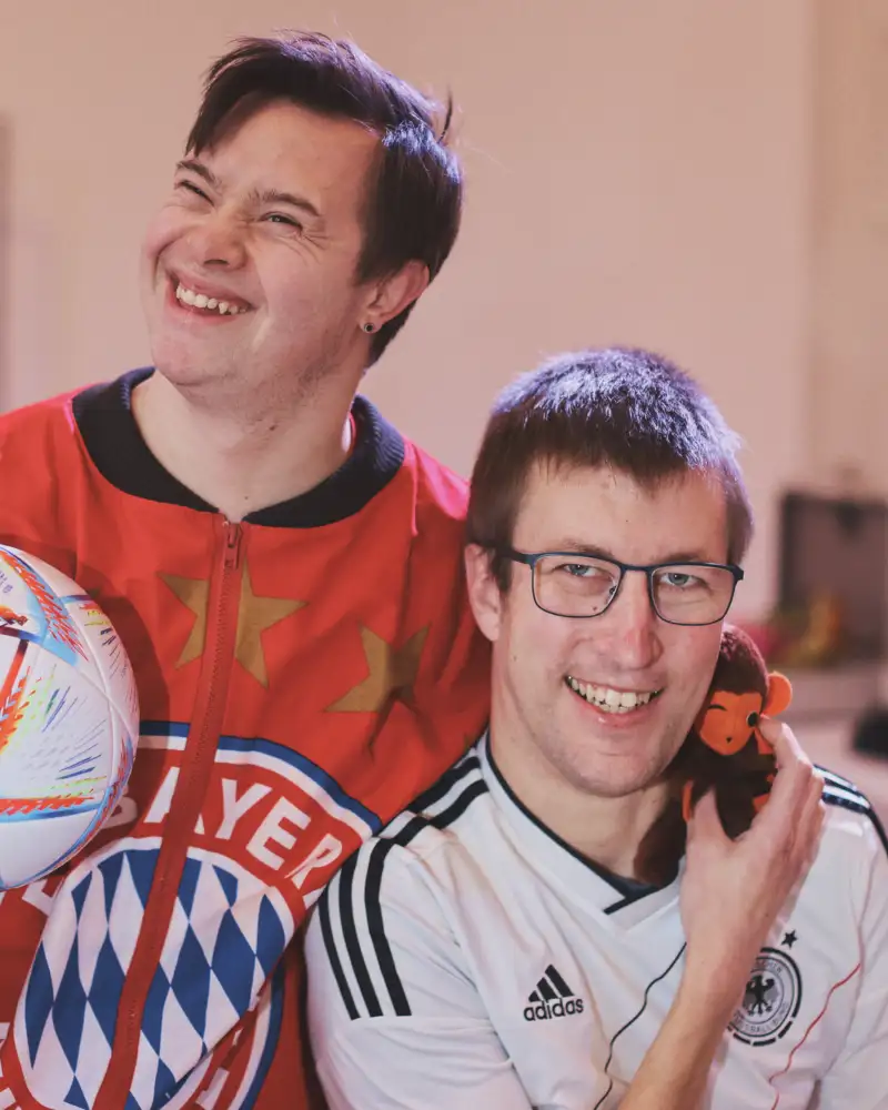 Zwei junge Männer in Sporttrikots stehen lachend nebeneinander. Der eine hält eine Fußball unter dem Arm.