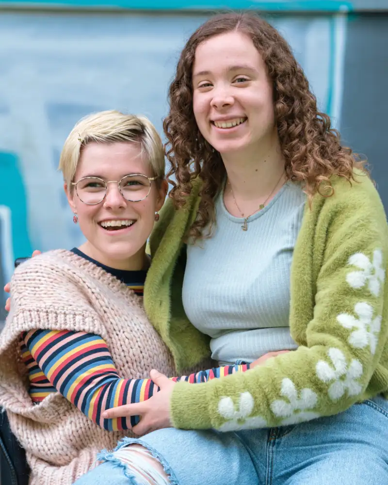 Zwei junge Frauen umarmen sich freundschaftlich. Eine von ihnen sitzt im Rollstuhl.