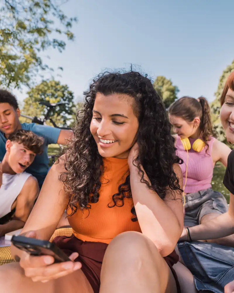 Eine Gruppe junger Menschen mit und ohne Behinderung sitzt in einem Park. Einige der Personen schauen auf ihre Smartphones.
