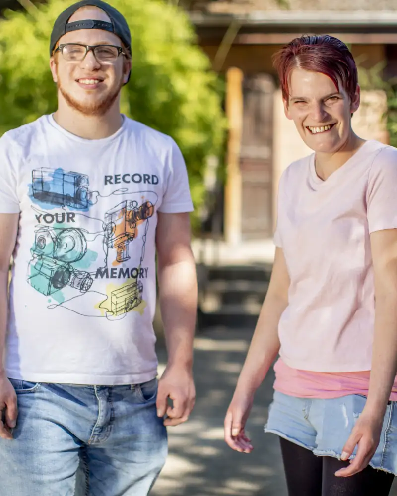 Okan und Zora. Zwei junge Menschen lachen in die Kamera. Okan hat einen rötlichen Bart und eine Brille. Er trägt ein nach hinten verdrehtes Basecap. Zora ist eine junge Frau mit kurzen, dunklen Haaren.