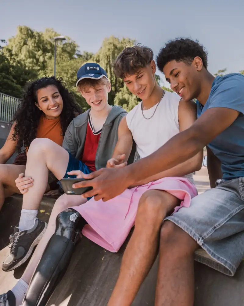 Vier Jugendliche unterschiedlicher Herkunft, mit und ohne Behinderung sitzen gemeinsam zusammen und schauen auf ein Telefon.