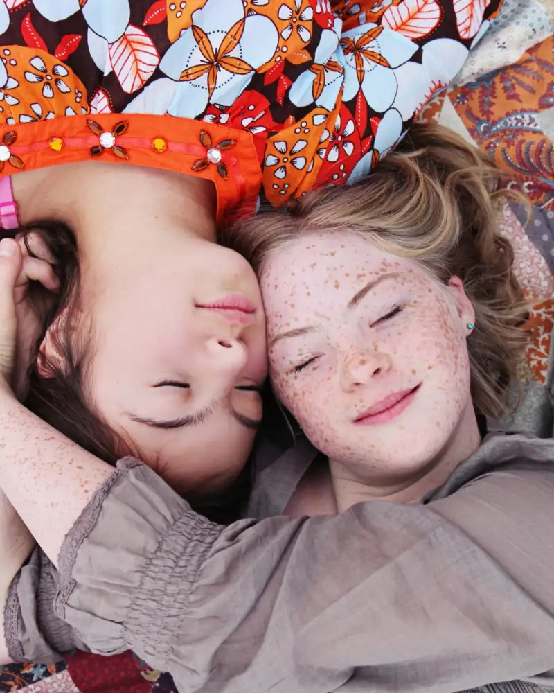 Blick von oben: zwei Mädchen liegen auf einer Decke mit ihren Köpfen dicht beieinander.