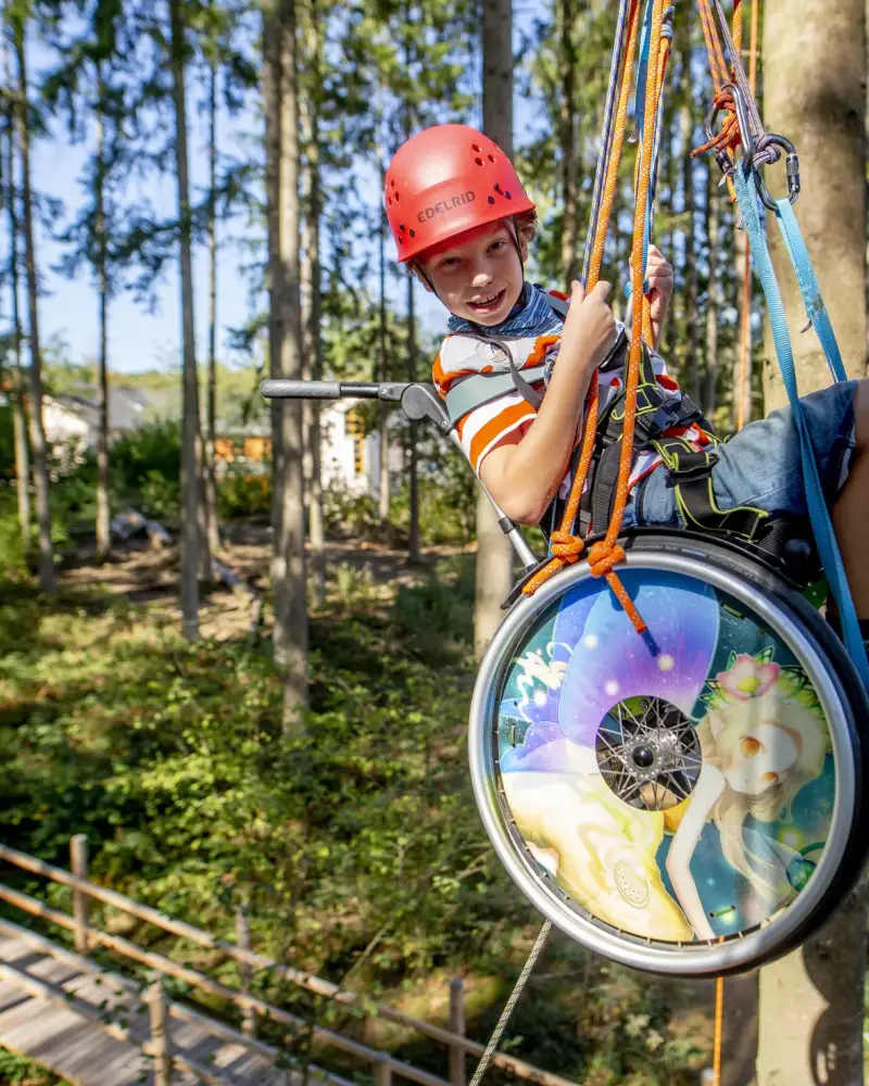 Ein Kind im Rollstuhl hängt an Seilen in einem Wald. Er trägt einen Schutzhelm und lacht in die Kamera.