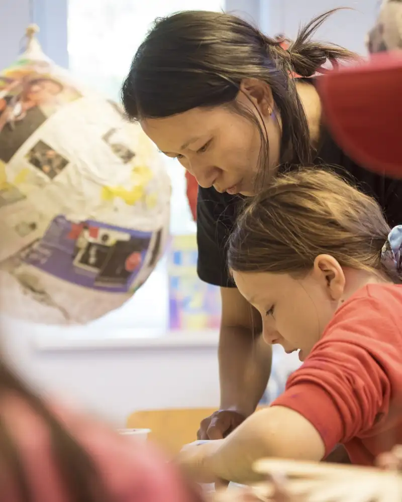 Ein Mädchen und eine junge Frau beugen sich gemeinsam über einen Tisch. Im Vordergrund sieht man unscharf zwei weitere Kinder. Im Hintergrund erkennt man ein Kunstobjekt.