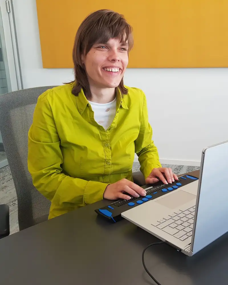 Eine blinde junge Frau sitzt lächelnd vor einem Laptop mit Braillezeile.