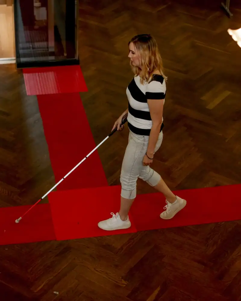 In einem großen Raum geht eine Frau mit einem Blindenlangstock an einer Markierung entlang.