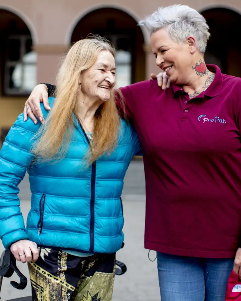 Eine Frau mit Rollator und eine weitere Frau liegen sich in den Armen und schauen sich an.