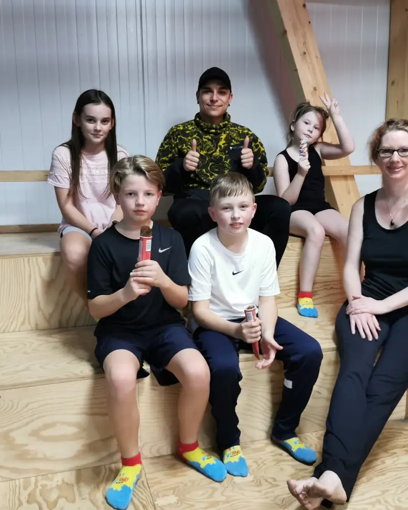 Eine Gruppe von vier Kindern, einem Jugendlichen und einer Frau sitzen auf Holzstufen. Alle tragen Turnkleidung.