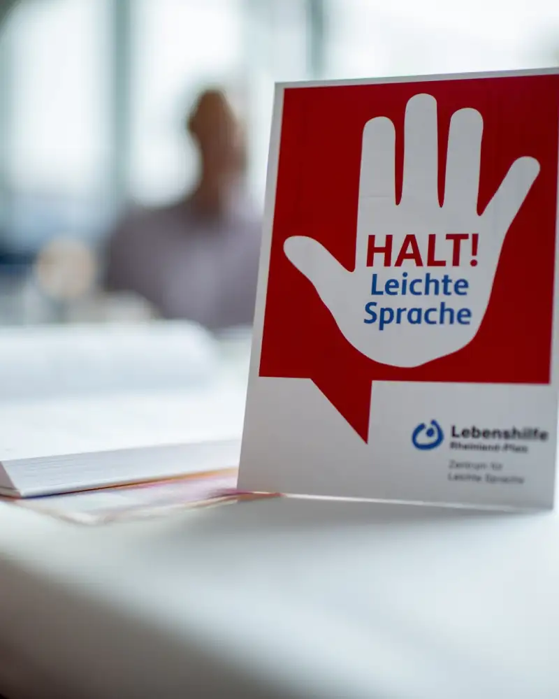 Auf einem Tisch steht ein Schild mit einer warnenden Hand und dem Hinweis: Halt! Leichte Sprache. Im Hintergrund sind unscharf drei Personen zu sehen.
