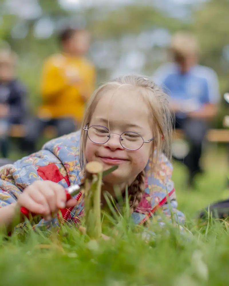 Auf einer Wiese liegt ein Mädchen mit Brille und schneidet eine Blume ab. Im Hintergrund kann man unscharf weitere Kinder sehen.