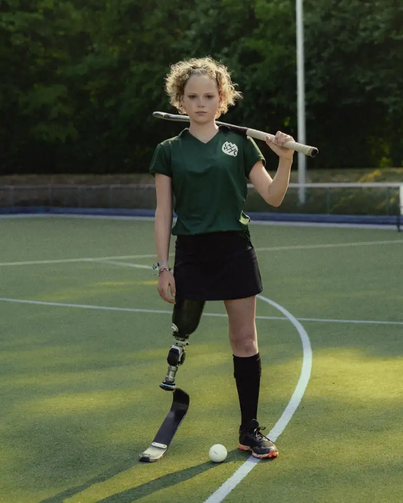 Annika, ein Mädchen mit lockigem Haar und Beinprothese steht auf einem Hockeyplatz. Zwischen ihren Beinen liegt der Ball, ihren Schläger hat sie lässig über die Schultern gelegt.
