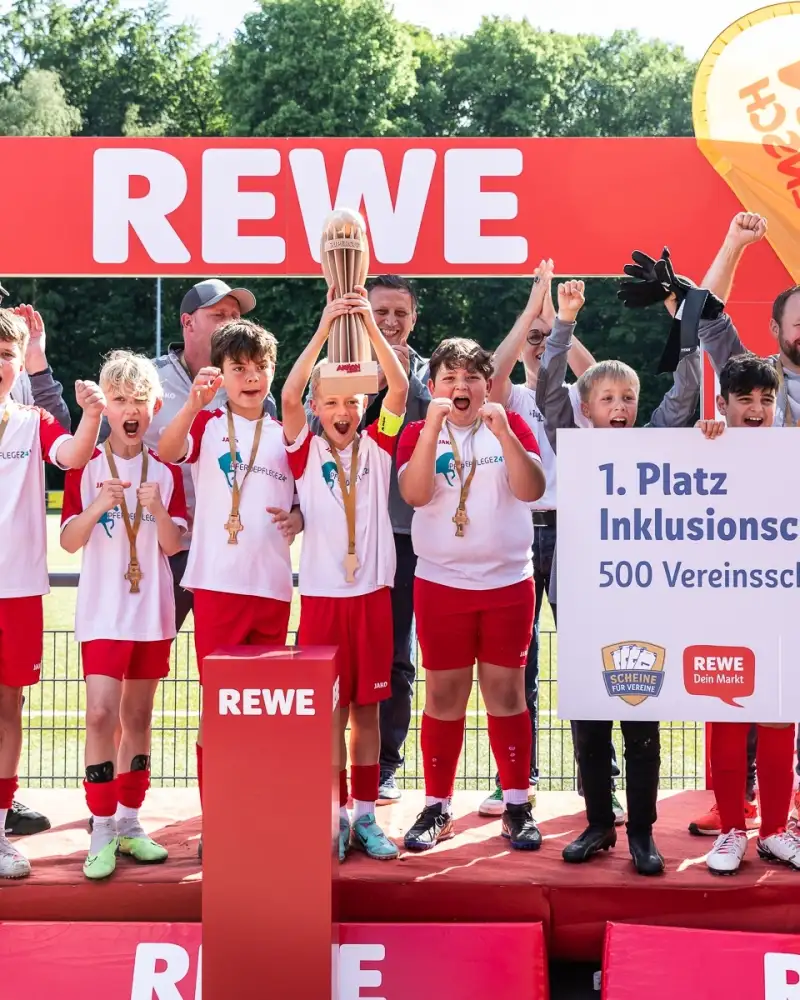 Eine Jungen-Fußballmannschaft feiert auf einem Podest bei der Siegerehrung ihren 1. Platz. Der Kapitän in der Mitte hält den Pokal in die Höhe, drei andere Jungen (etwa 12 Jahre) halten ein Schild in den Händen auf dem steht: "1. Platz Inklusionscup - 500 Vereinsscheine".