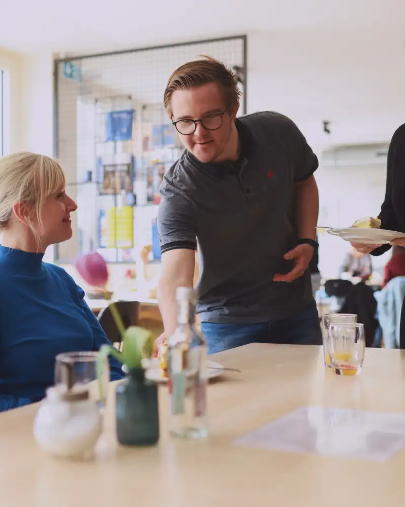 Zwei Frauen sitzen an einem Holztisch. Ein Mann mit Down-Syndrom und eine junge Frau servieren jeder einen Teller mit Kuchen.