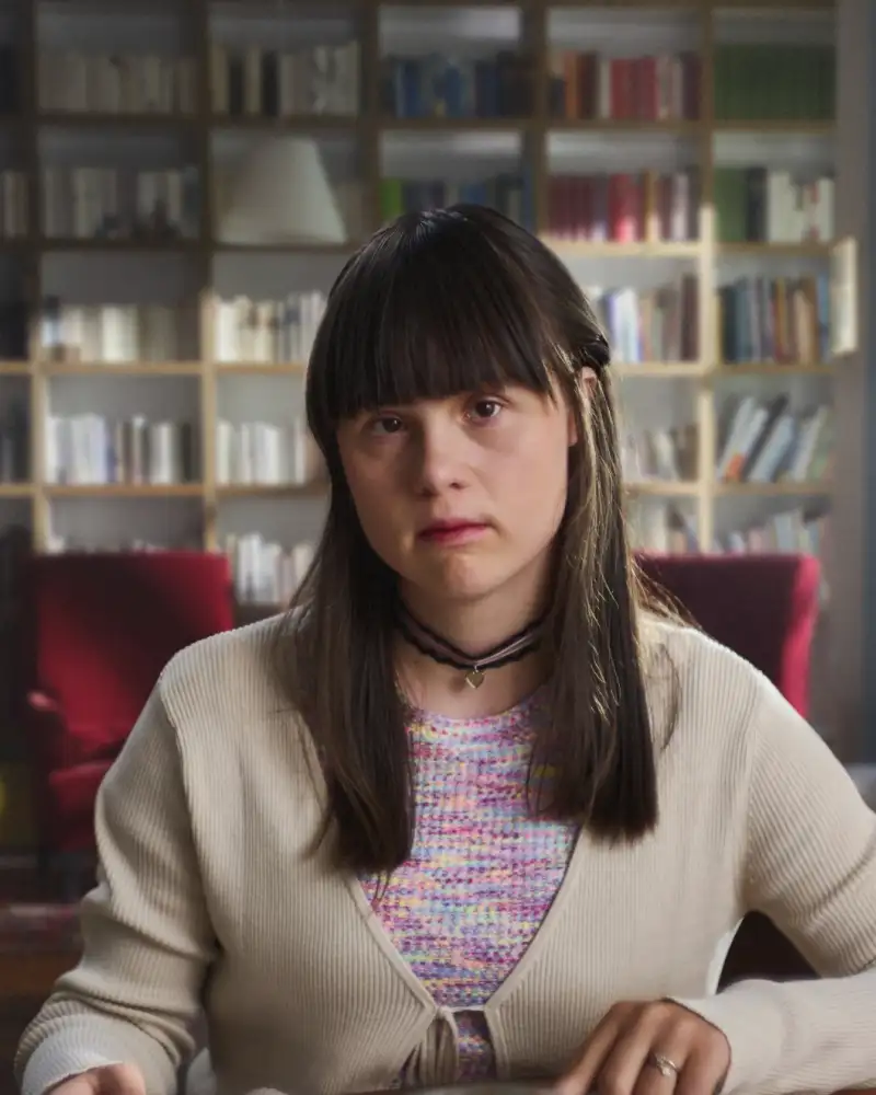 Eine junge Frau mit langen dunklen Haaren schaut bekümmert in die Kamera. Im Hintergrund sieht man zwei Sessel und ein Bücherregal.